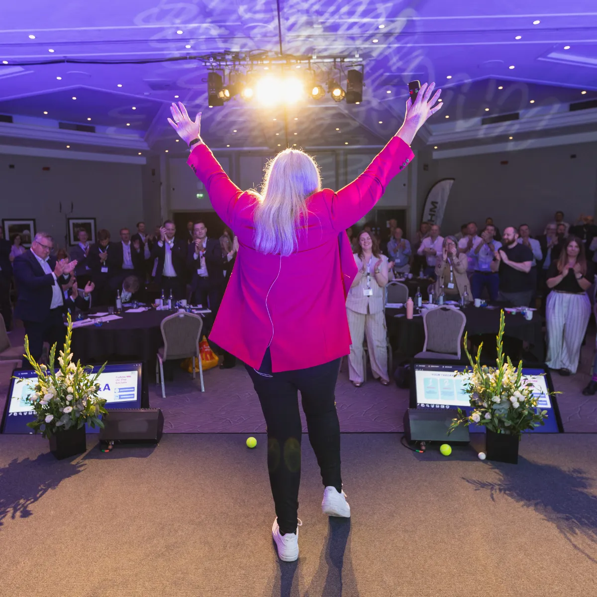 Dr Alison Edgar MBE, arms aloft on stage, as the audience rises in a standing ovation.