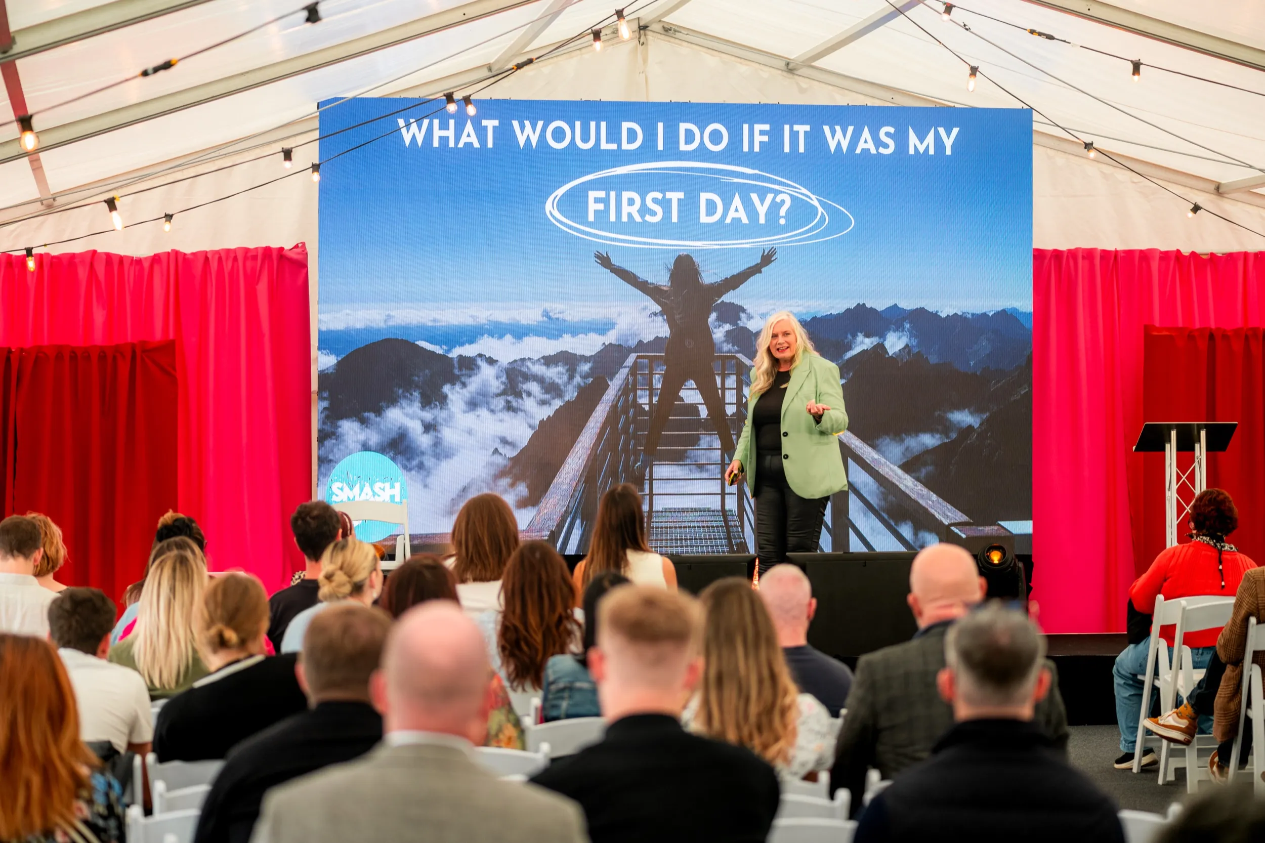 Alison on stage at Bizzfest, the words 'What would I do if it was my first day?' filling the screen behind her.