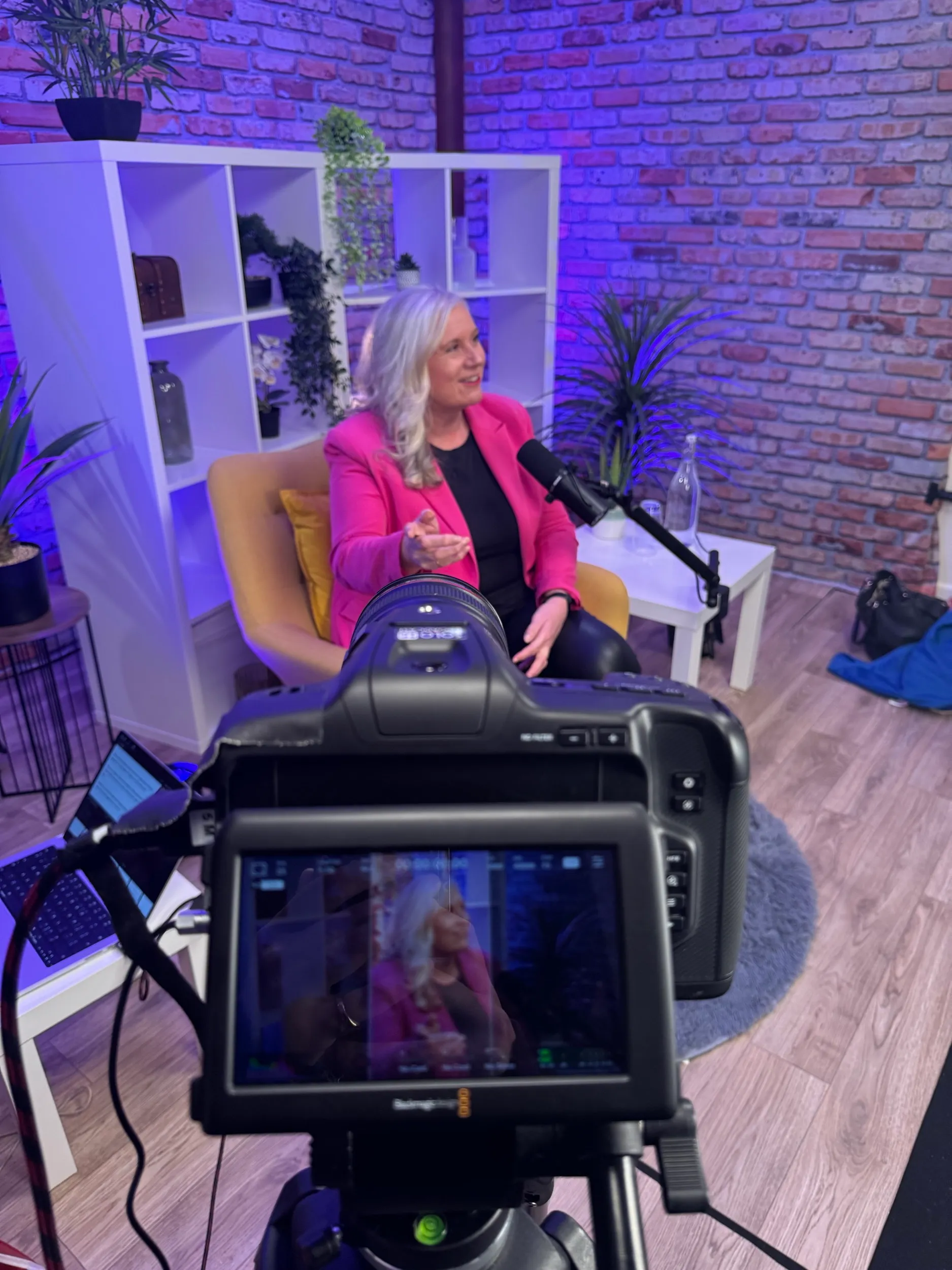 Alison at the microphone in a podcast studio, mid-thought in a pink blazer.